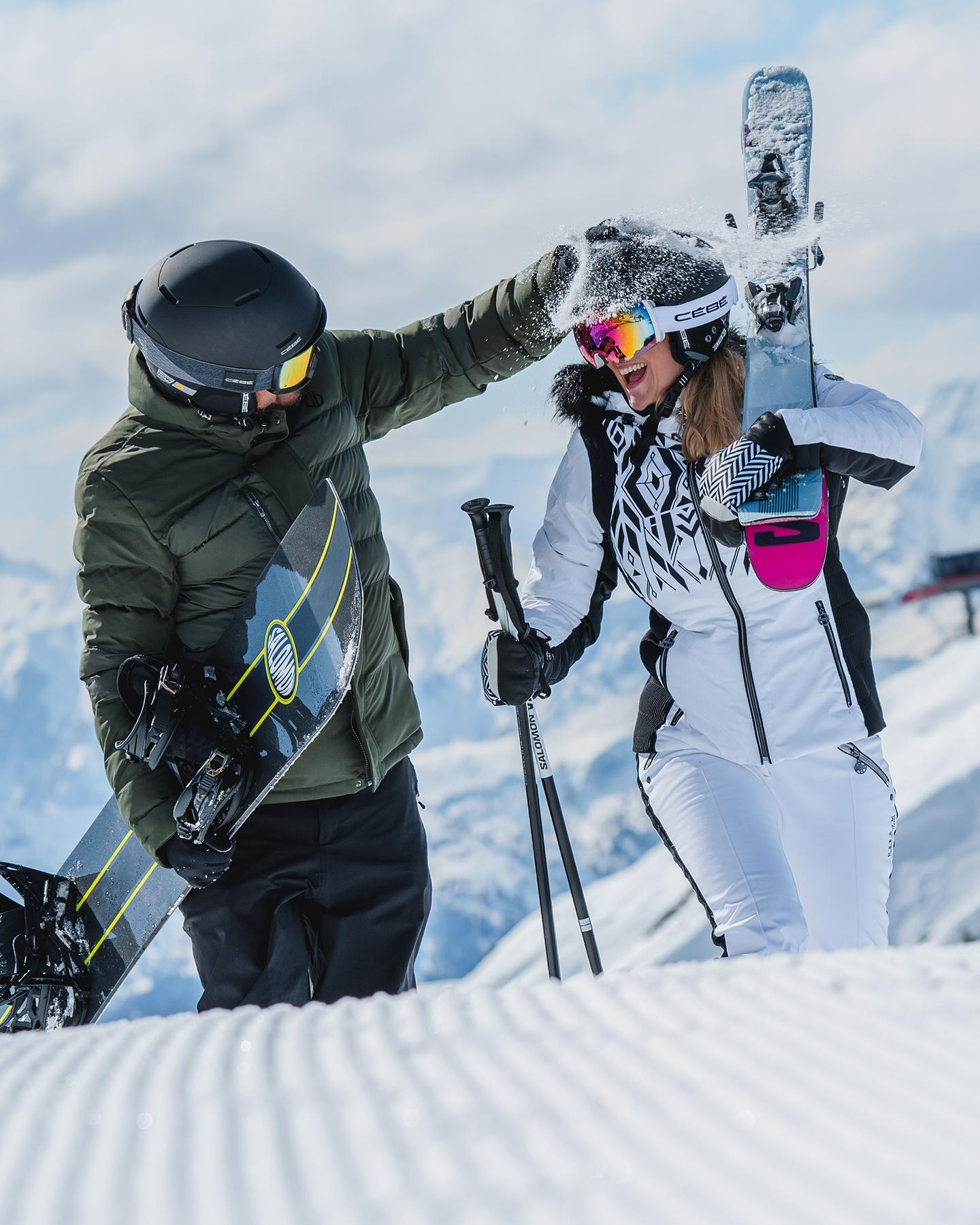 Un couple de personnes marchent en s'amusant avec la neige sur une piste de Paradiski avec leurs skis et snowboard à la main 